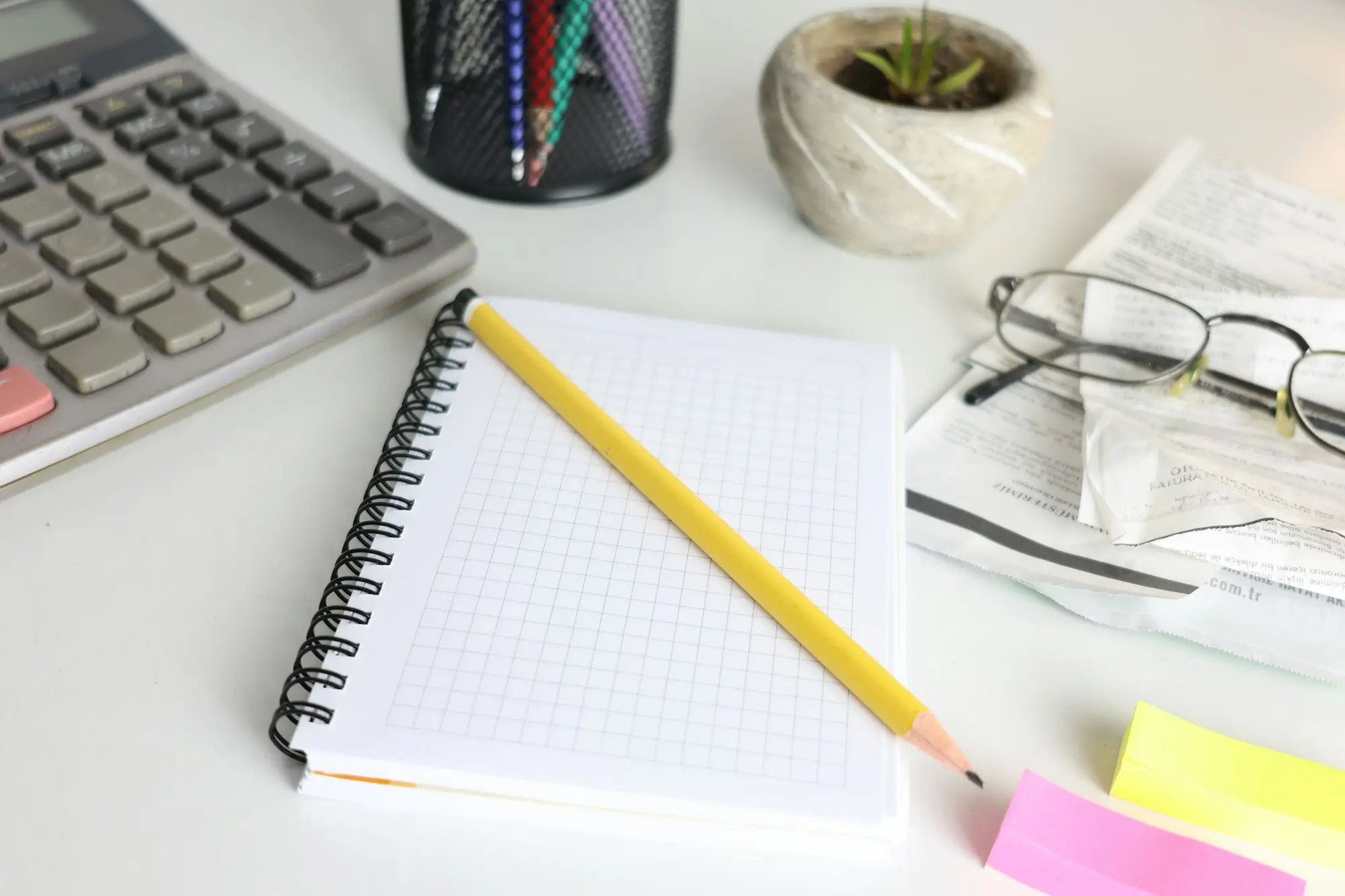 A desk with a calculator, spiral notepad, pencil, glasses and printed receipts laid out for working out costs.