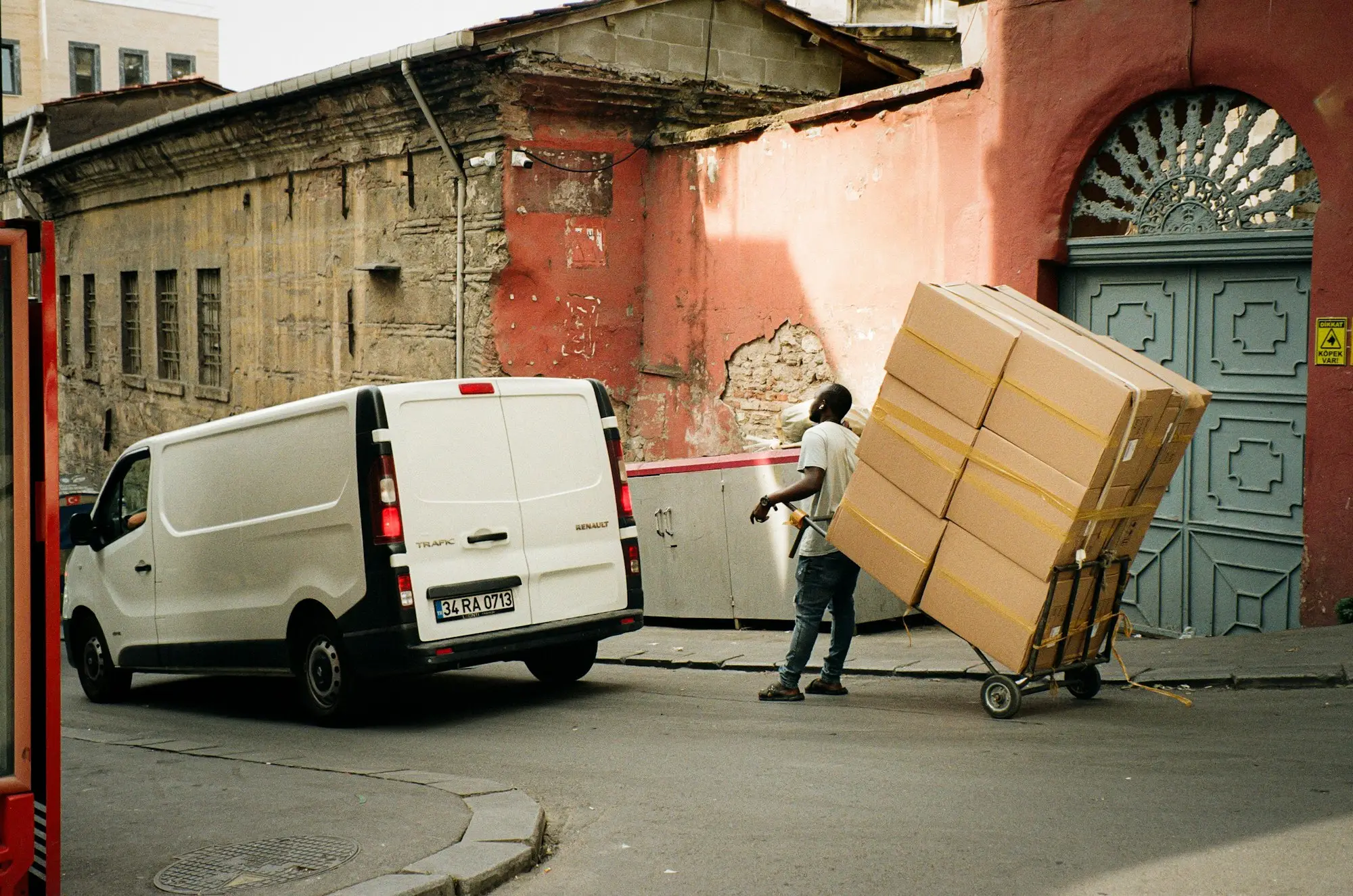 A delivery driver pulling a trolley stacked with cardboard boxes beside a white van on a quiet street in daylight.