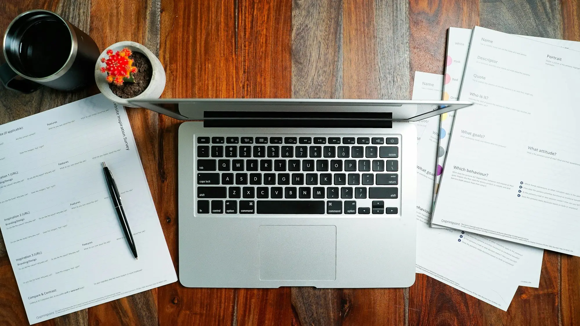 A laptop on a timber desk surrounded by printed statements, a pen and a coffee.