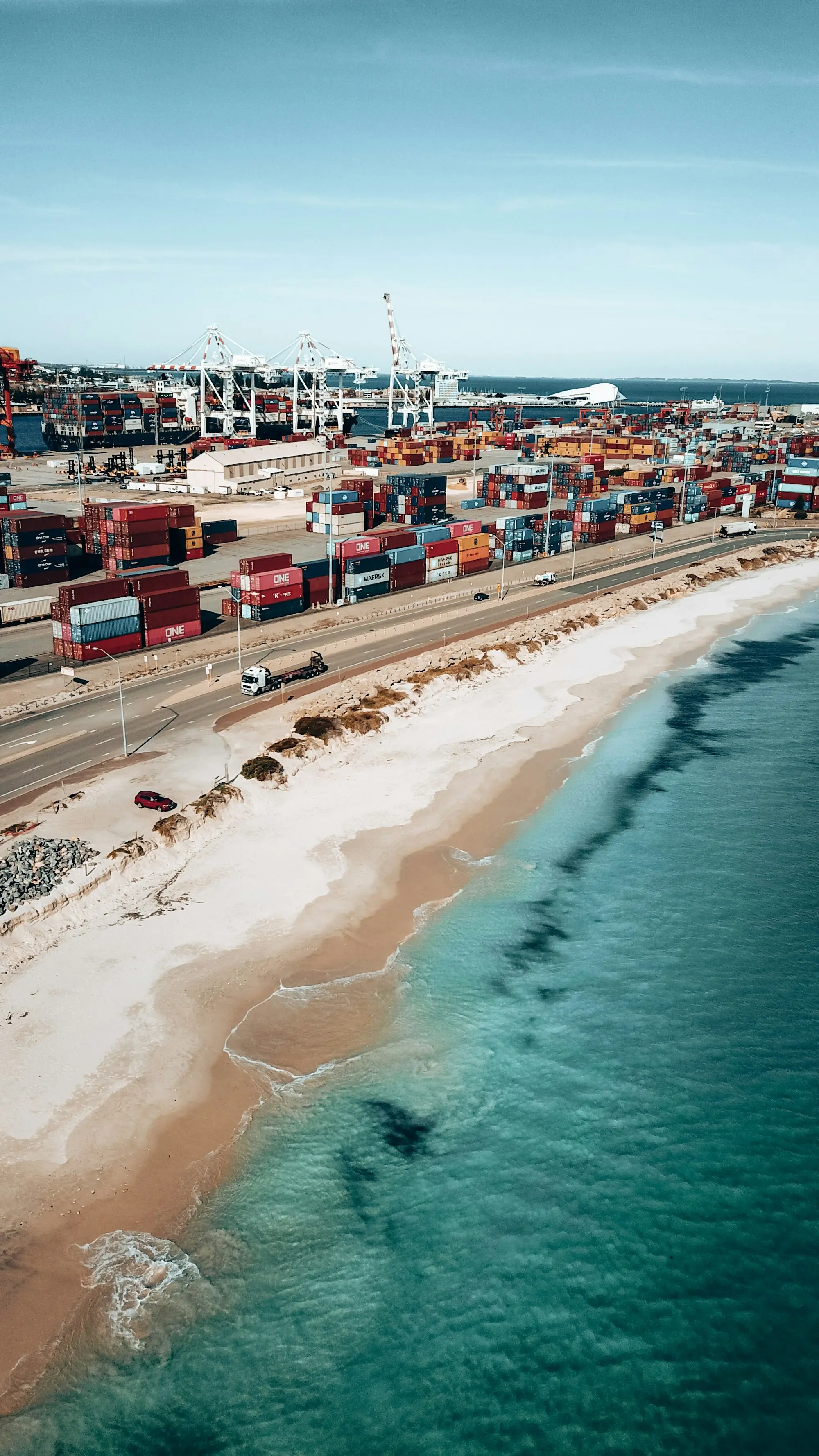Aerial view of stacked shipping containers and port cranes beside a sandy beach and turquoise water.