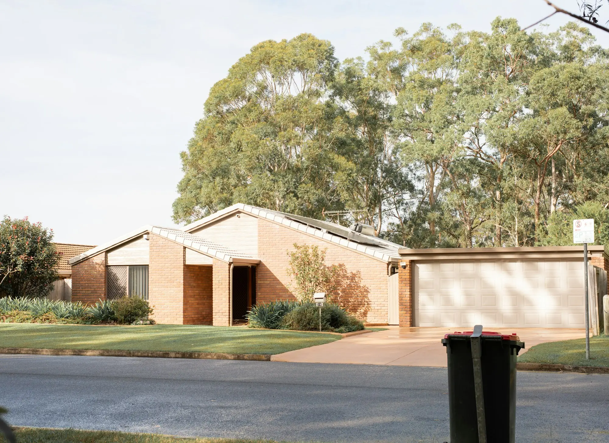 A single-storey brick house with a front garden, double garage and wheelie bin on a leafy suburban street.