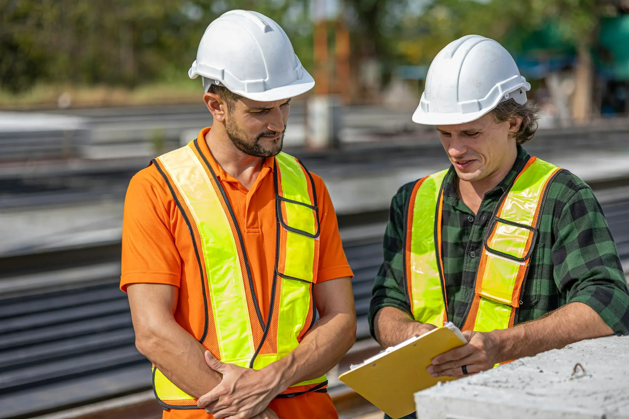 Two tradesmen in hard hats and hi-vis vests reviewing paperwork on a clipboard at a work site.