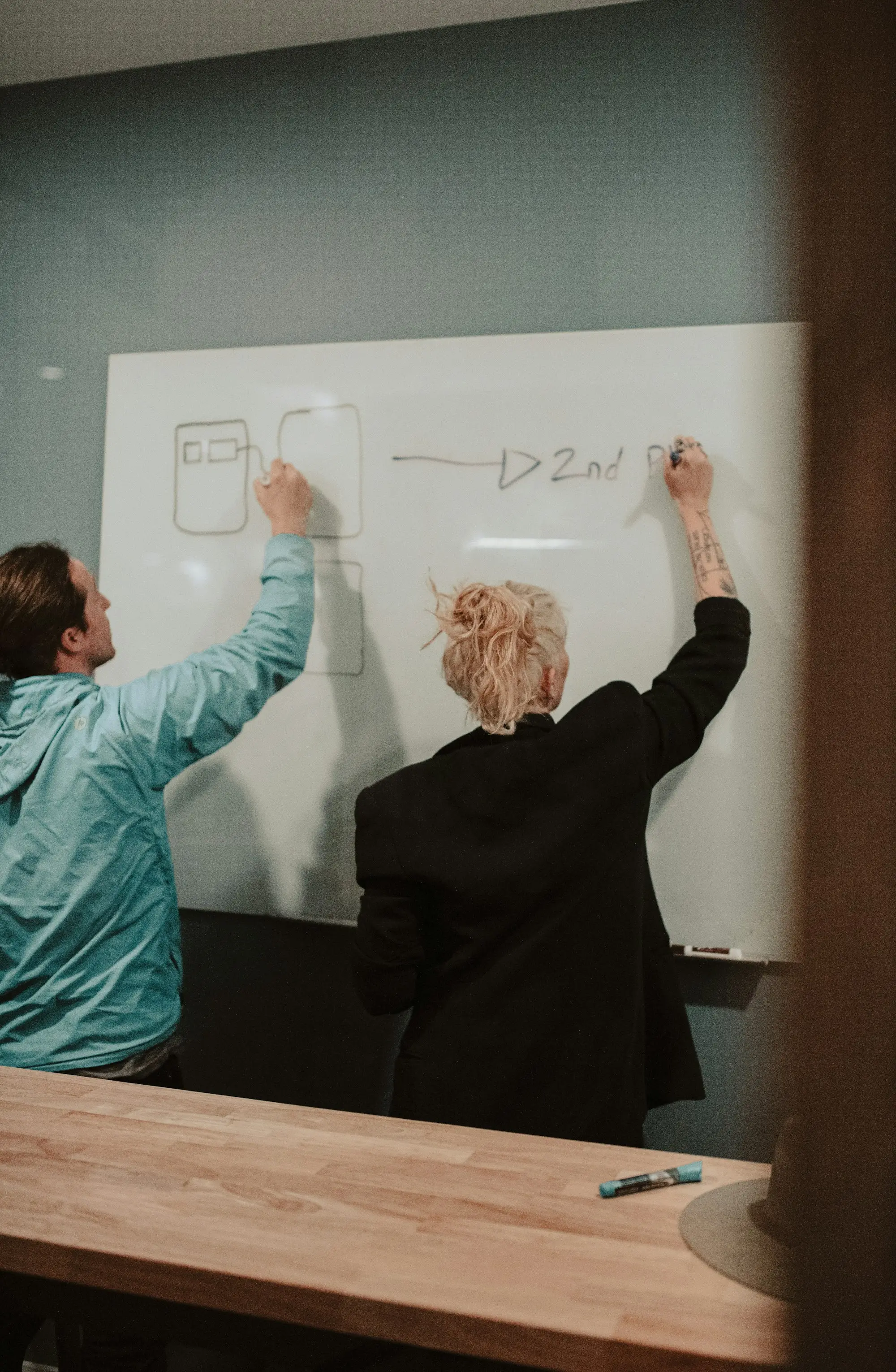 Two people drawing boxes and arrows on a whiteboard in an office meeting room.
