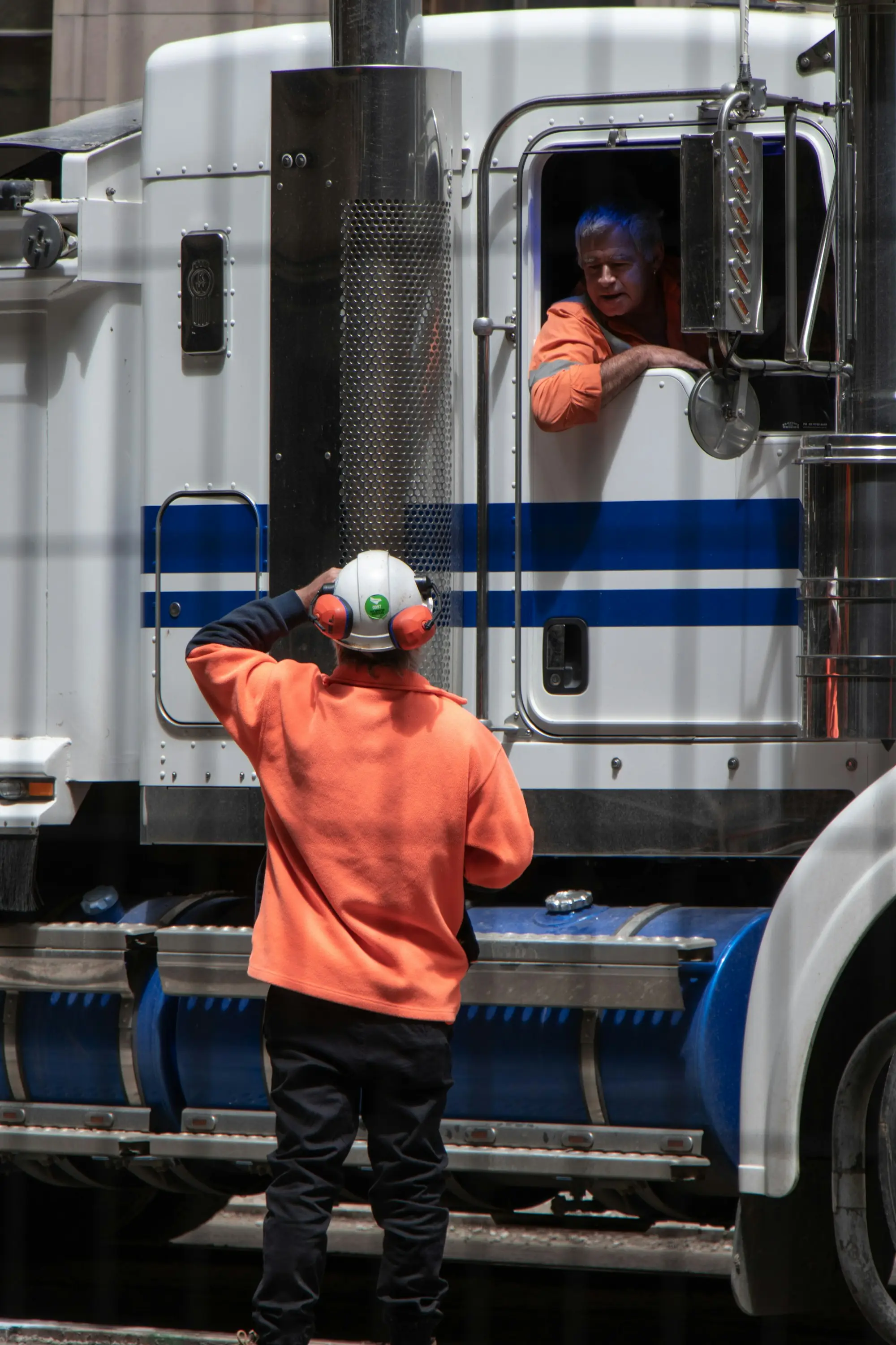 A worker in a hi-vis shirt and hard hat inspecting a heavy truck while the driver looks on from the cab.