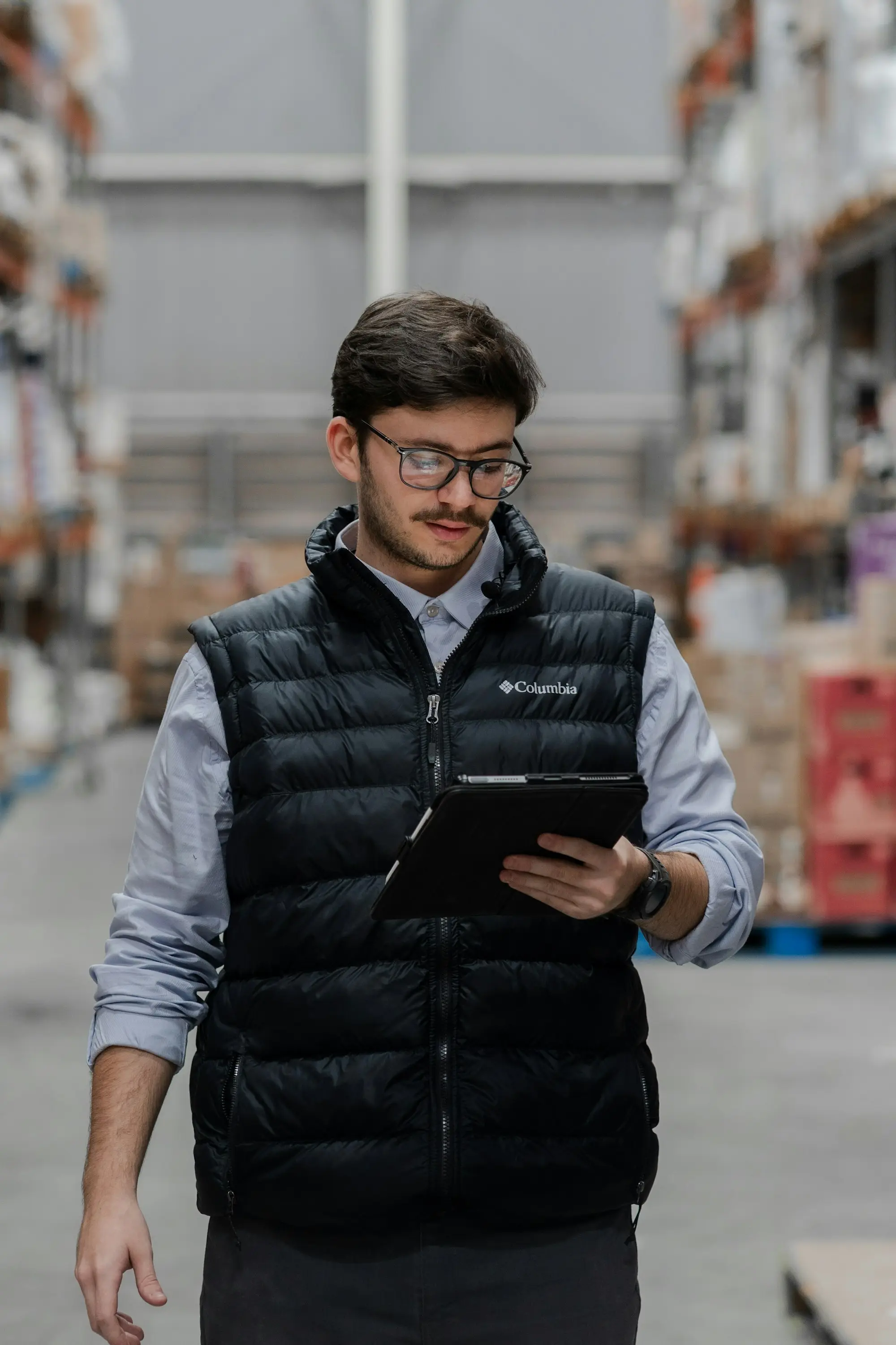A man in a vest checking stock on a tablet while walking down a warehouse aisle lined with racking.