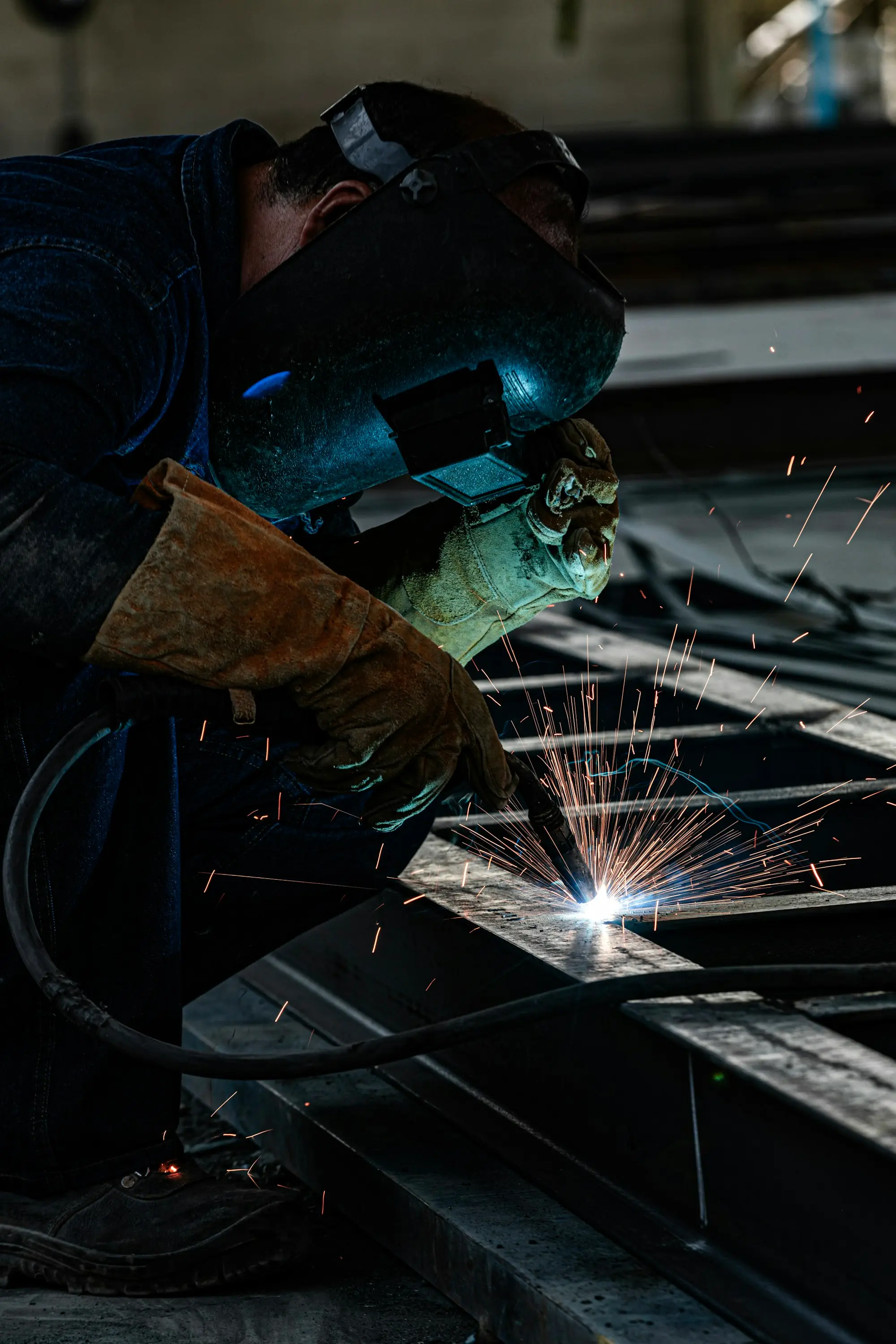 A welder in a mask and heavy gloves working on steel with sparks flying in a workshop.